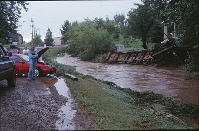 A flooded creek with stopped and stalled cars next to it on a dirt road, with a washed up dock flattened on the opposite shore